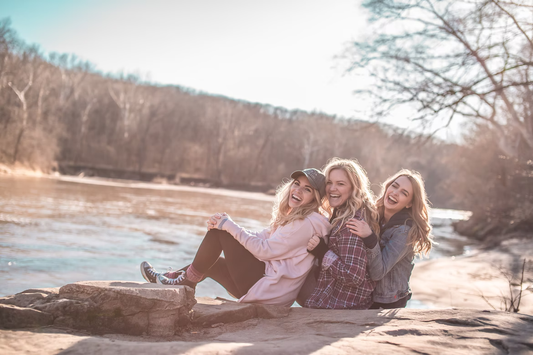 Drei Freundinnen sitzen lachend nebeneinander am sonnigen Fluss.