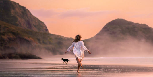 Frau rennt mit Hund über nassen Strand im Sonnenuntergang.