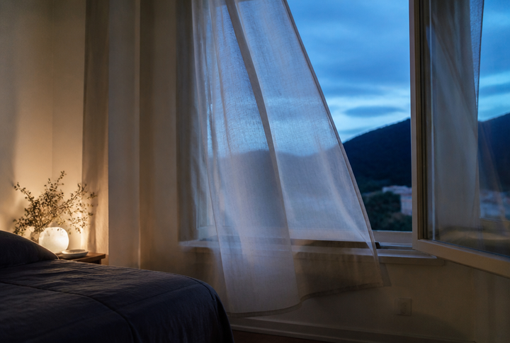 Vorhang weht vor einem offenen Schlafzimmerfenster zur blauen Stunde